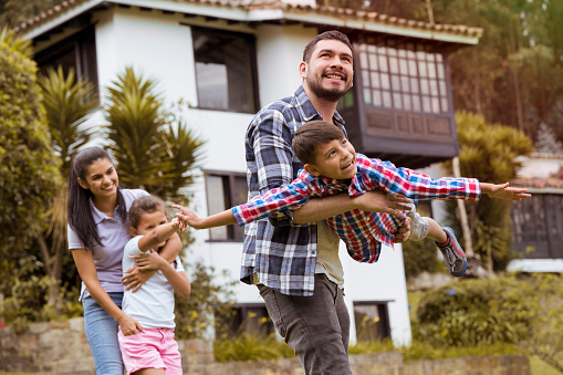 Beautiful family, enjoying an afternoon outside their house, the father and the child are playing the plane, smiling happily, while the mom and the girl are watching the others play, happy